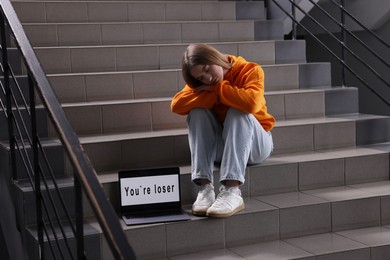 Photo of Cyber bullying. Upset teenage girl and laptop with text You Loser on stairs indoors