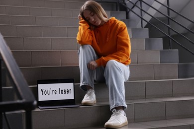 Photo of Cyber bullying. Upset teenage girl and laptop with text You Loser on stairs indoors