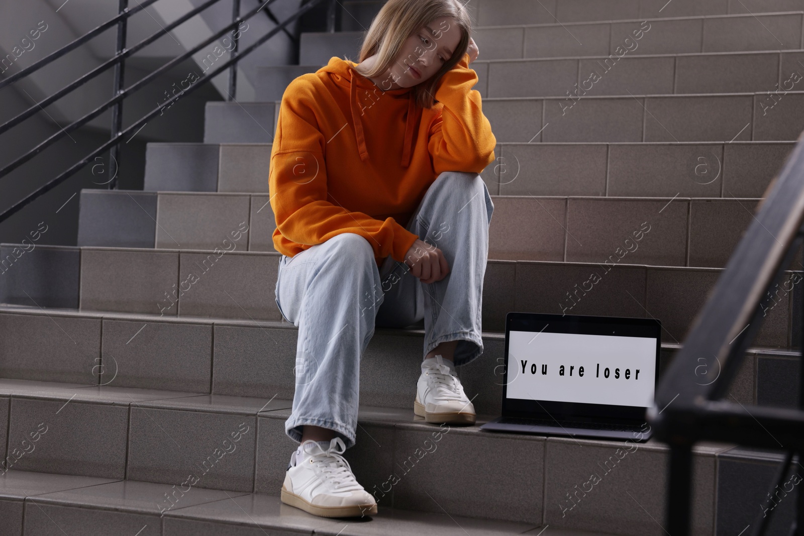 Cyber bullying. Upset teenage girl and laptop with text You Loser on stairs indoors Photo of Cyber bullying. Upset teenage girl and laptop with text You Loser on stairs indoors