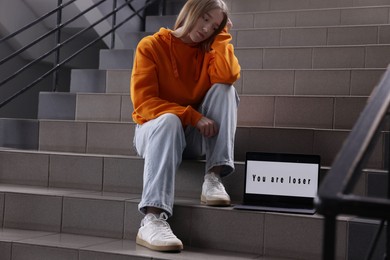 Photo of Cyber bullying. Upset teenage girl and laptop with text You Loser on stairs indoors
