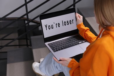 Photo of Cyber bullying. Upset teenage girl looking at laptop with text You Loser on stairs indoors, closeup