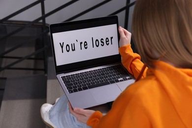 Photo of Cyber bullying. Upset teenage girl looking at laptop with text You Loser on stairs indoors, closeup