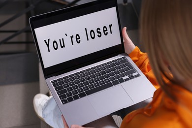 Photo of Cyber bullying. Upset teenage girl looking at laptop with text You Loser on stairs indoors, closeup