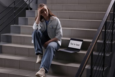 Photo of Cyber bullying. Upset teenage girl and laptop with text You Loser on stairs indoors