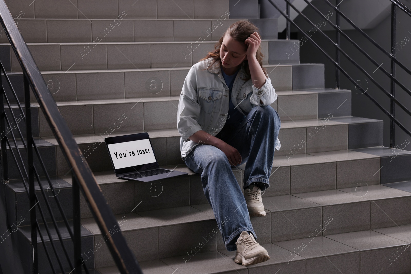 Cyber bullying. Upset teenage girl and laptop with text You Loser on stairs indoors Photo of Cyber bullying. Upset teenage girl and laptop with text You Loser on stairs indoors