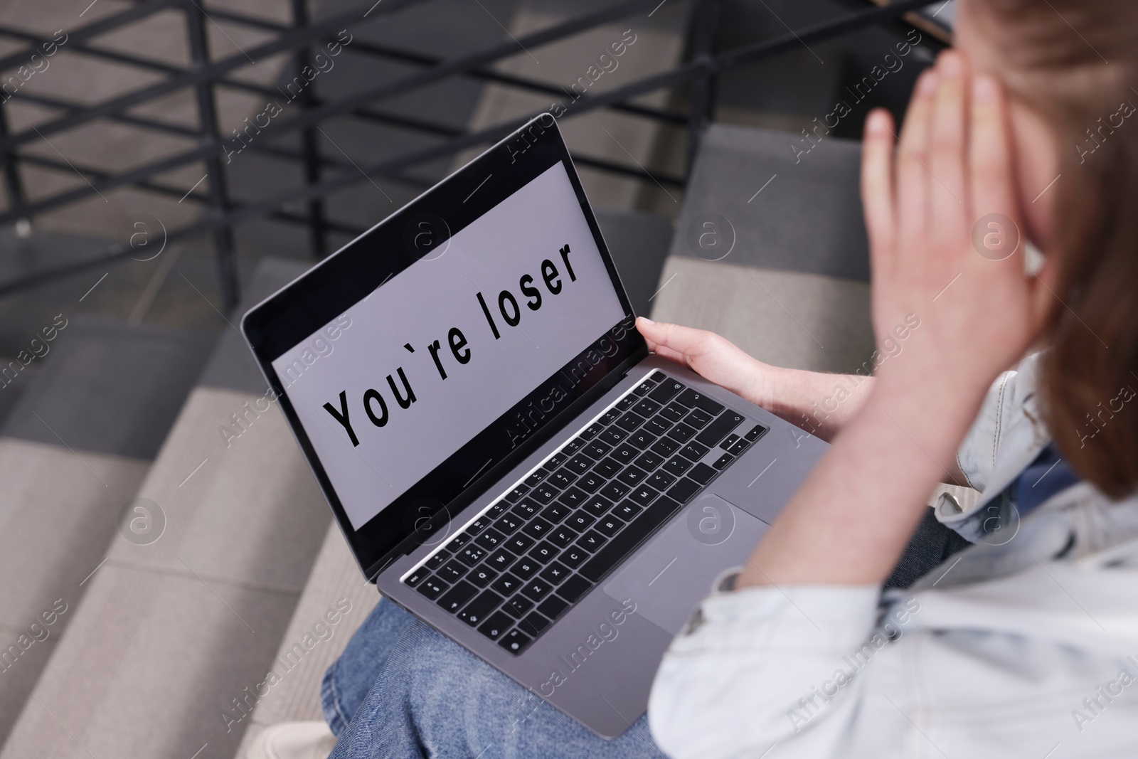 Cyber bullying. Upset teenage girl looking at laptop with text You Loser on stairs indoors, closeup Photo of Cyber bullying. Upset teenage girl looking at laptop with text You Loser on stairs indoors, closeup