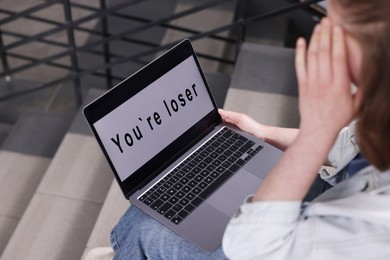 Photo of Cyber bullying. Upset teenage girl looking at laptop with text You Loser on stairs indoors, closeup