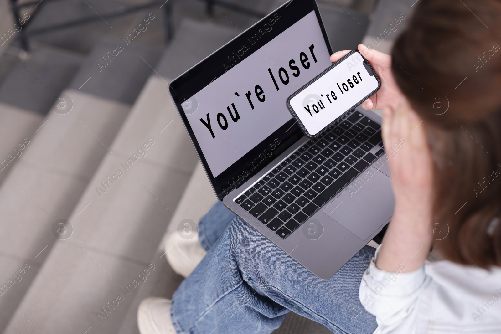 Cyber bullying. Upset teenage girl looking at laptop and smartphone with text You Loser on stairs indoors, closeup Photo of Cyber bullying. Upset teenage girl looking at laptop and smartphone with text You Loser on stairs indoors, closeup