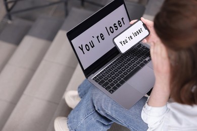 Photo of Cyber bullying. Upset teenage girl looking at laptop and smartphone with text You Loser on stairs indoors, closeup