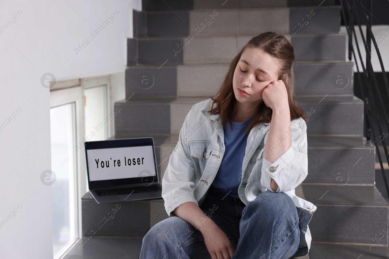 Cyber bullying. Upset teenage girl and laptop with text You Loser on stairs indoors Photo of Cyber bullying. Upset teenage girl and laptop with text You Loser on stairs indoors