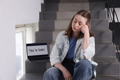 Photo of Cyber bullying. Upset teenage girl and laptop with text You Loser on stairs indoors