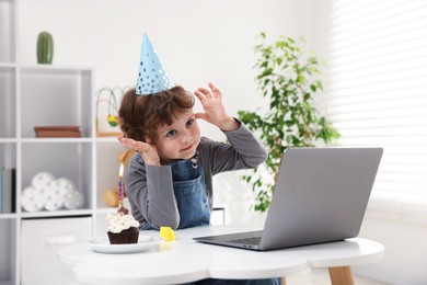 Photo of Little boy celebrating his birthday while having video call via laptop at table indoors