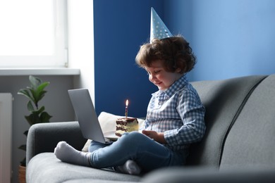 Photo of Birthday celebration. Little boy with piece of cake having video call via laptop on sofa at home