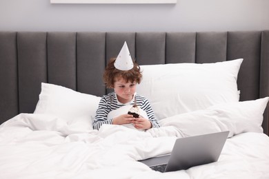 Photo of Birthday celebration. Little boy with cupcake having video call via laptop in bed at home