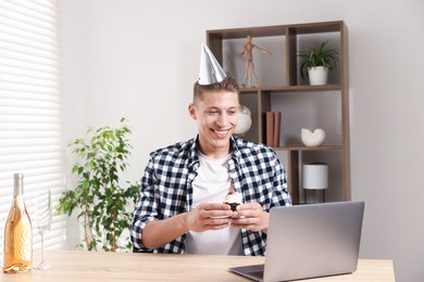 Photo of Birthday celebration. Man with cupcake and sparkling wine having video call via laptop at table indoors