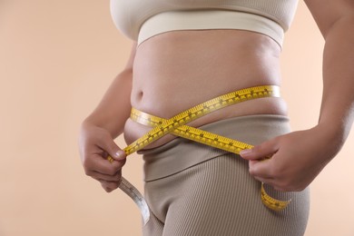 Photo of Overweight woman measuring her belly with tape on beige background, closeup
