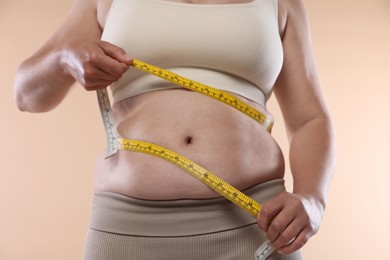 Photo of Overweight woman measuring her belly with tape on beige background, closeup