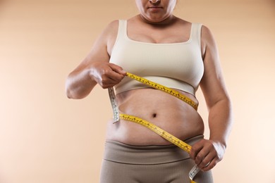 Photo of Overweight woman measuring her belly with tape on beige background, closeup