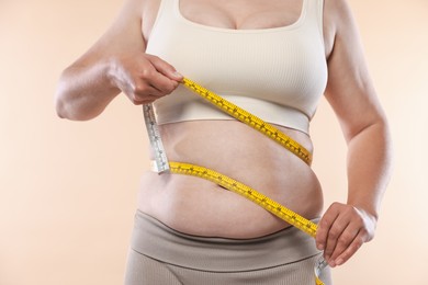 Photo of Overweight woman measuring her belly with tape on beige background, closeup