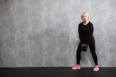 Photo of Senior woman with kettlebell near gray marble wall indoors, space for text