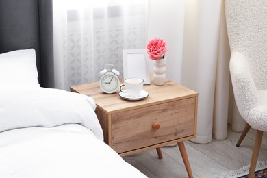 Photo of Bedside table with coffee, flower and alarm clock near bed indoors
