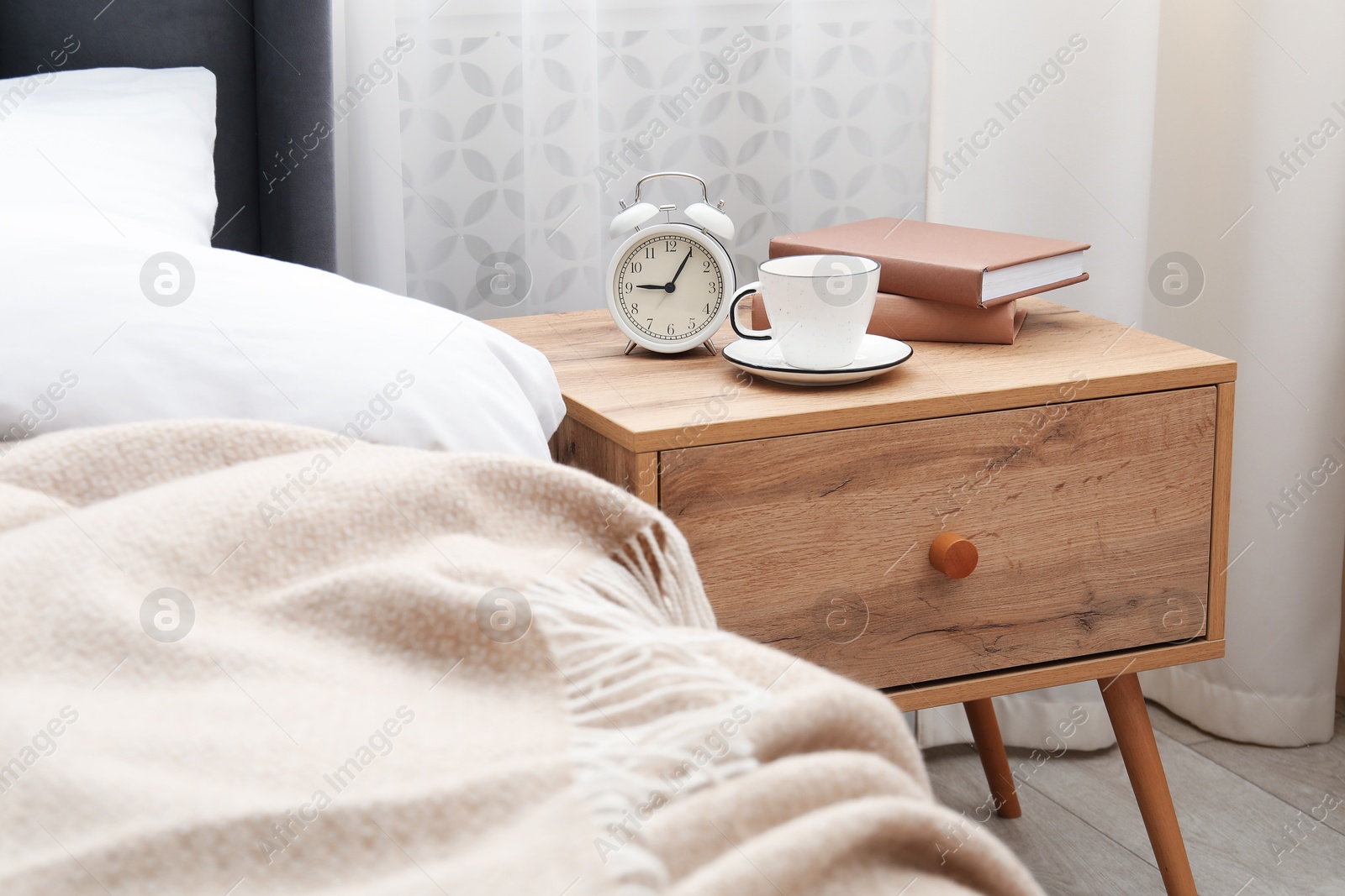 Bedside table with coffee, books and alarm clock near bed indoors Photo of Bedside table with coffee, books and alarm clock near bed indoors