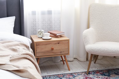 Photo of Bedside table with coffee, books and alarm clock near bed indoors