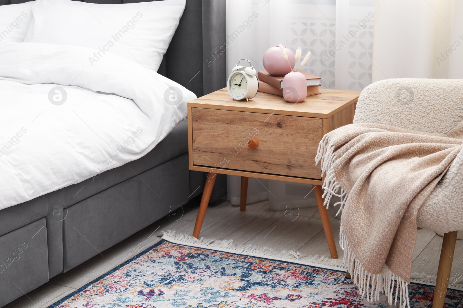 Bedside table with alarm clock and decor near bed indoors Photo of Bedside table with alarm clock and decor near bed indoors
