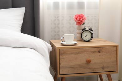 Photo of Bedside table with coffee, flower and alarm clock near bed indoors