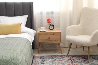 Photo of Bedside table with coffee, flower and alarm clock near bed indoors