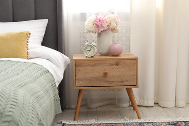 Photo of Bedside table with alarm clock and flowers near bed indoors
