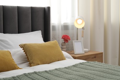 Photo of Bedside table with lamp, flower and frame near bed indoors