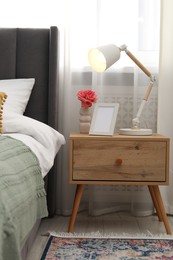 Photo of Bedside table with lamp, flower and frame near bed indoors
