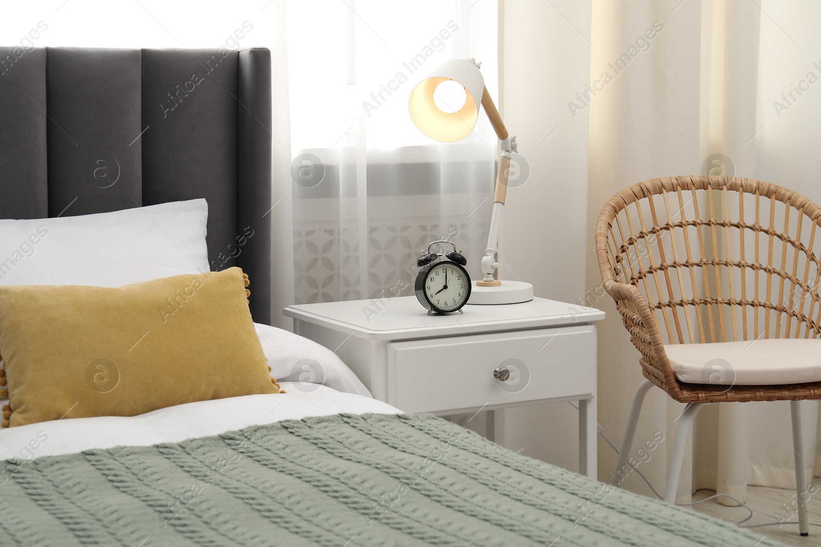 Bedside table with lamp and alarm clock near bed indoors Photo of Bedside table with lamp and alarm clock near bed indoors