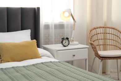 Photo of Bedside table with lamp and alarm clock near bed indoors
