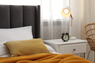Photo of Bedside table with lamp and alarm clock near bed indoors