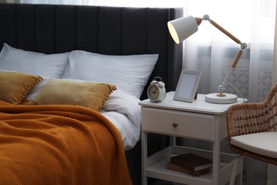 Photo of Bedside table with lamp, frame and alarm clock near bed indoors