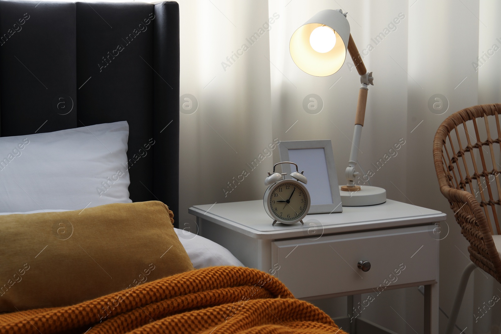 Bedside table with lamp, frame and alarm clock near bed indoors Photo of Bedside table with lamp, frame and alarm clock near bed indoors