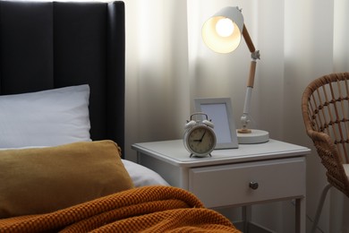 Photo of Bedside table with lamp, frame and alarm clock near bed indoors
