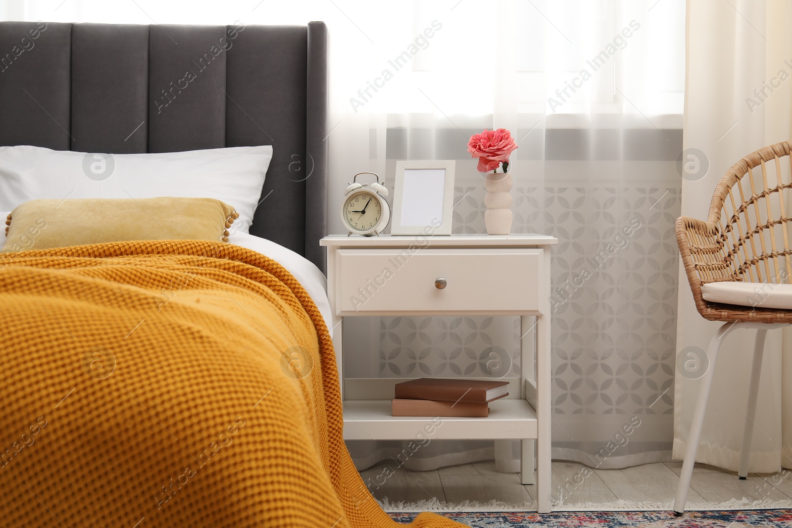 Bedside table with alarm clock, flower and frame near bed indoors Photo of Bedside table with alarm clock, flower and frame near bed indoors