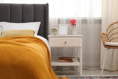 Photo of Bedside table with alarm clock, flower and frame near bed indoors