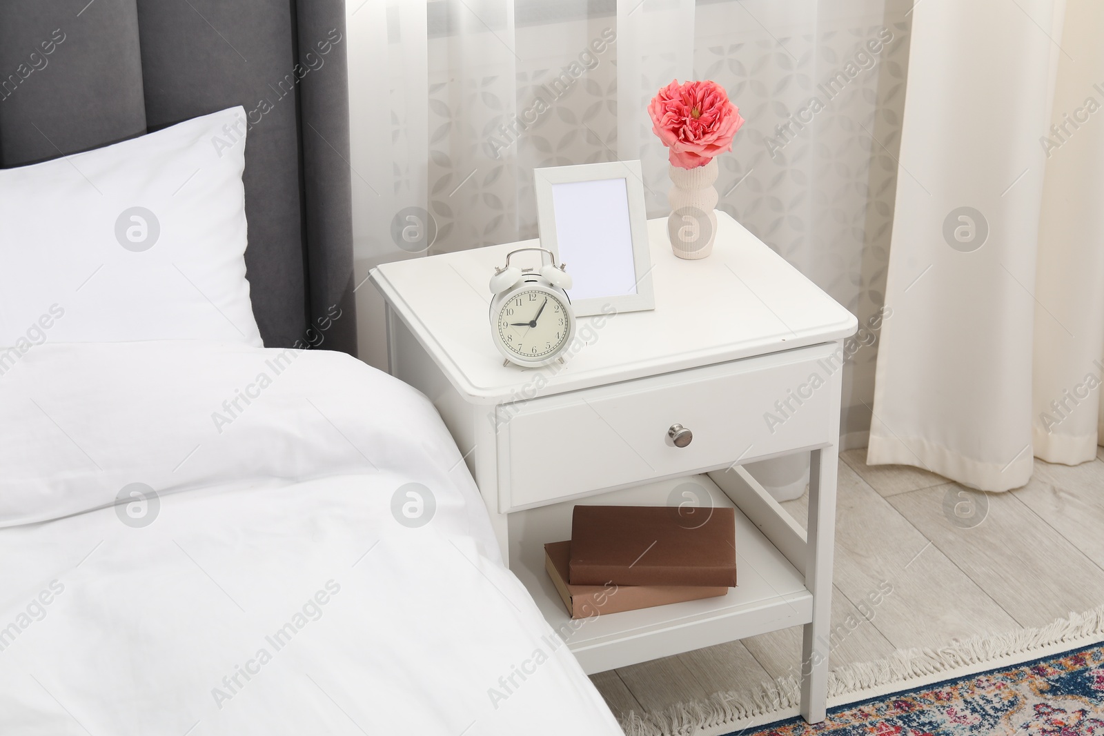 Bedside table with alarm clock, flower and frame near bed indoors Photo of Bedside table with alarm clock, flower and frame near bed indoors
