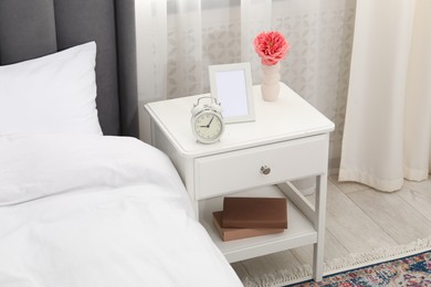 Photo of Bedside table with alarm clock, flower and frame near bed indoors