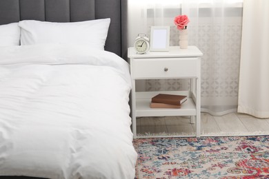 Photo of Bedside table with alarm clock, flower and frame near bed indoors