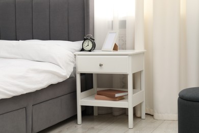 Photo of Bedside table with alarm clock and frame near bed indoors