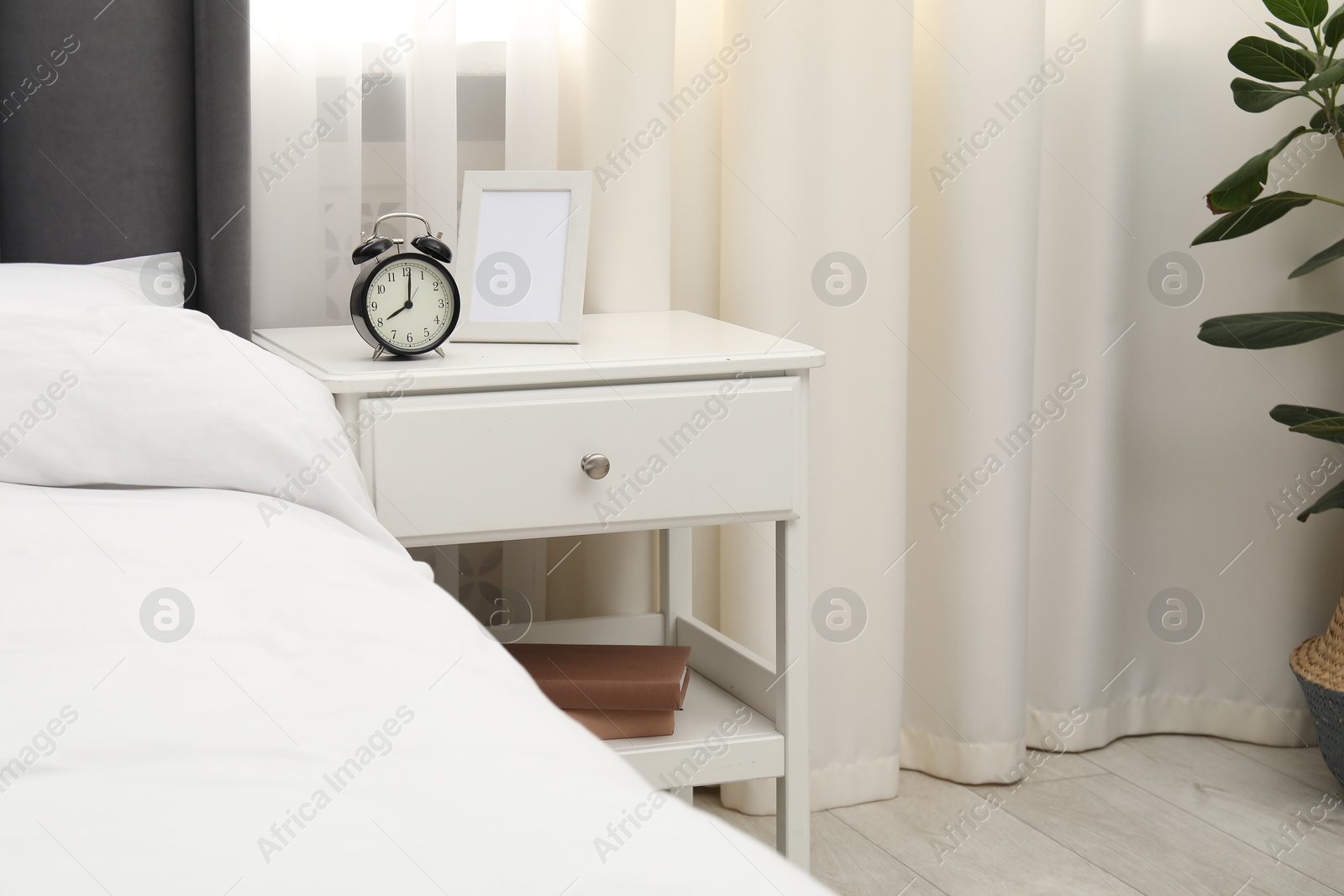Bedside table with alarm clock and frame near bed indoors Photo of Bedside table with alarm clock and frame near bed indoors