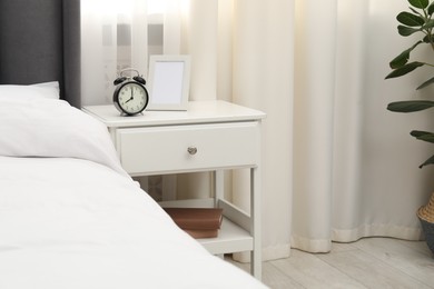 Photo of Bedside table with alarm clock and frame near bed indoors