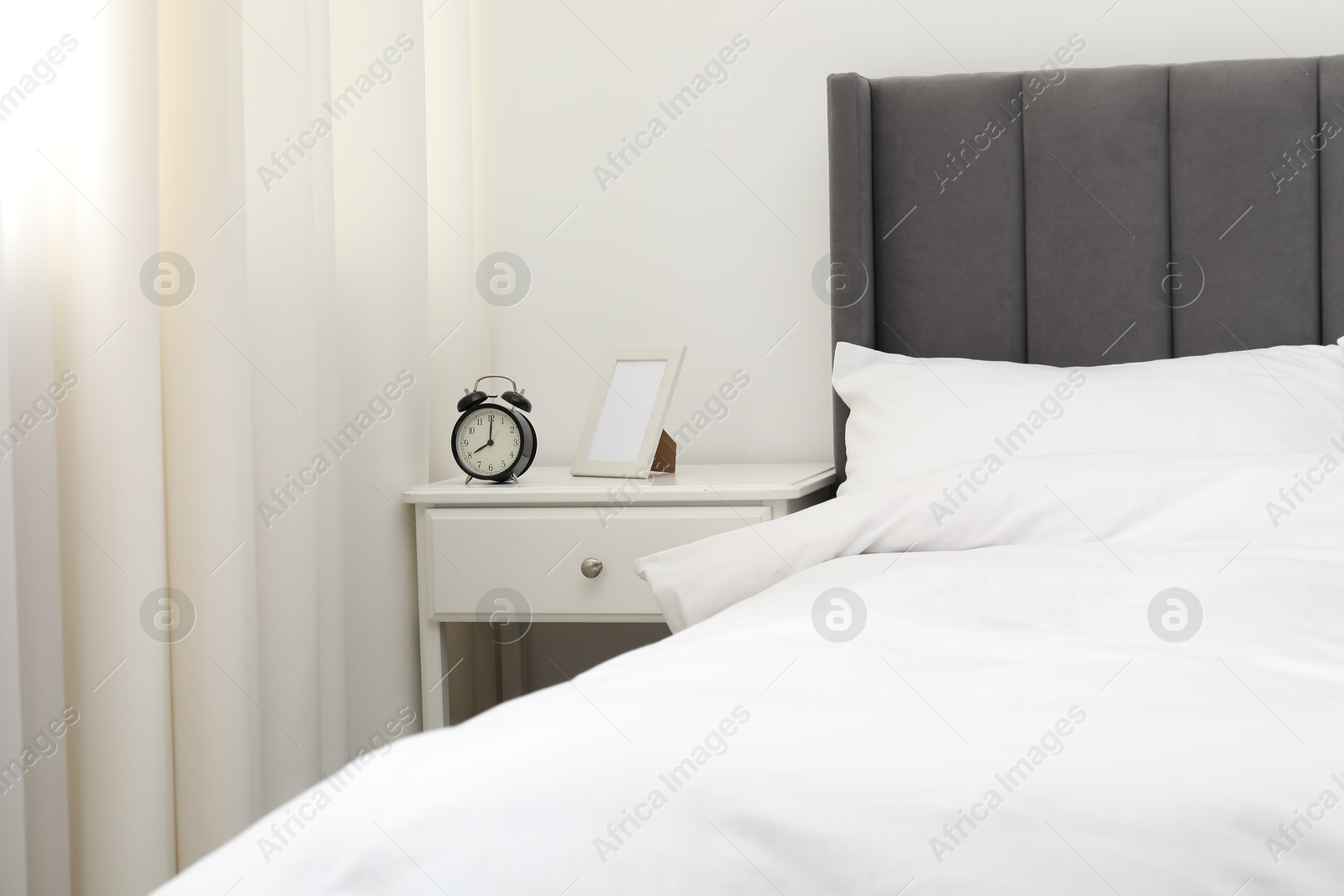 Bedside table with alarm clock and frame near bed indoors Photo of Bedside table with alarm clock and frame near bed indoors