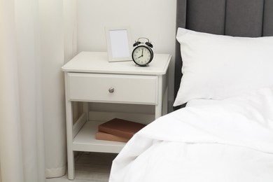 Photo of Bedside table with alarm clock and frame near bed indoors