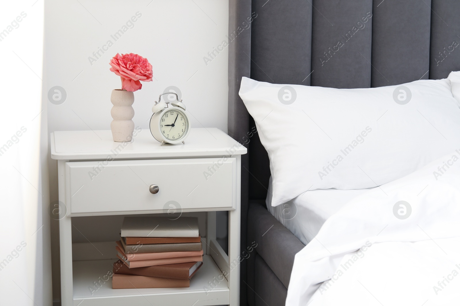 Bedside table with alarm clock and flower near bed indoors Photo of Bedside table with alarm clock and flower near bed indoors